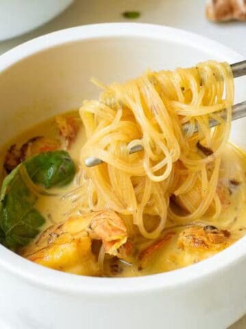 A bowl of noodle soup with pieces of chicken, basil leaves, and broth, with chopsticks lifting noodles from the bowl.