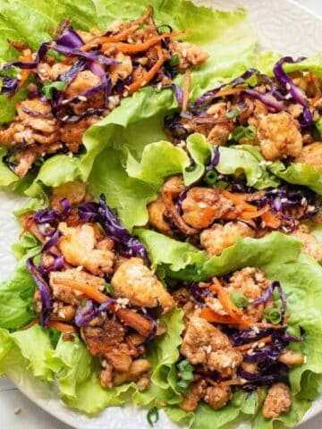 Four lettuce wraps filled with cooked chicken, shredded carrots, and purple cabbage are arranged on a white plate, with a small bowl of shredded cabbage and sesame seeds visible nearby.