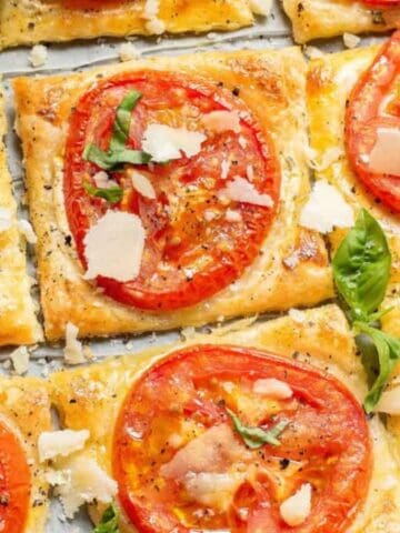 Squares of tomato tart made with puff pastry, topped with sliced tomatoes, cheese shavings, black pepper, and fresh basil, arranged on parchment paper.