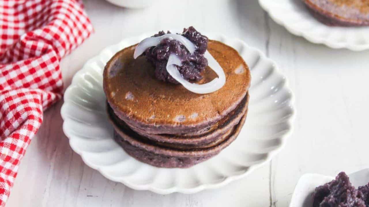 A stack of three purple pancakes topped with sweet purple paste and white strips, served on a white plate with a red checkered cloth nearby.
