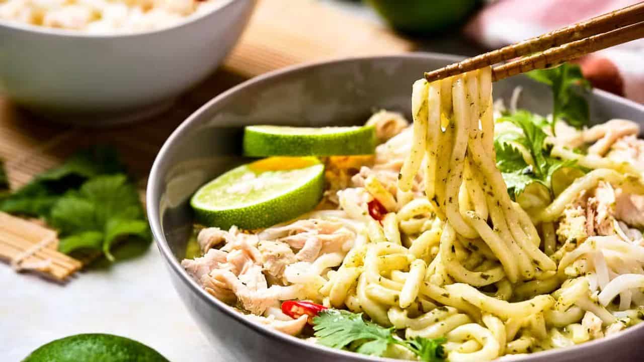 A bowl of green curry noodles topped with shredded chicken, lime slices, red chili slices, and fresh cilantro. Chopsticks are lifting a portion of the noodles from the bowl. A soft background showcases another bowl and fresh herbs.