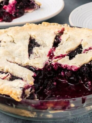 A baked blueberry pie with a golden crust, several slices removed, revealing the filling. Two plates with pie slices are in the background.