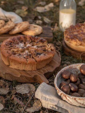 A picnic scene with pies, cookies, a bowl of chestnuts, a glass bottle of milk, and a cloth napkin arranged on grass with fallen leaves.