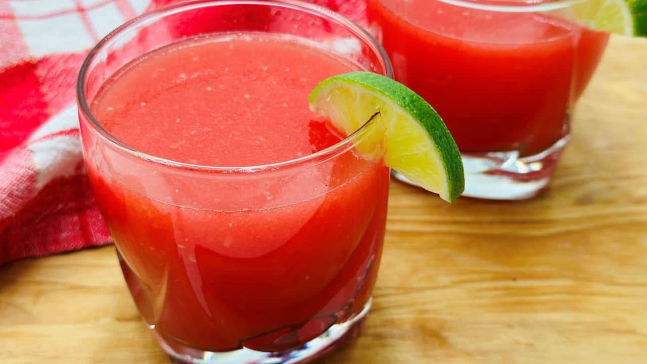 Two glasses of watermelon juice garnished with lime slices are on a wooden surface with a red napkin in the background.