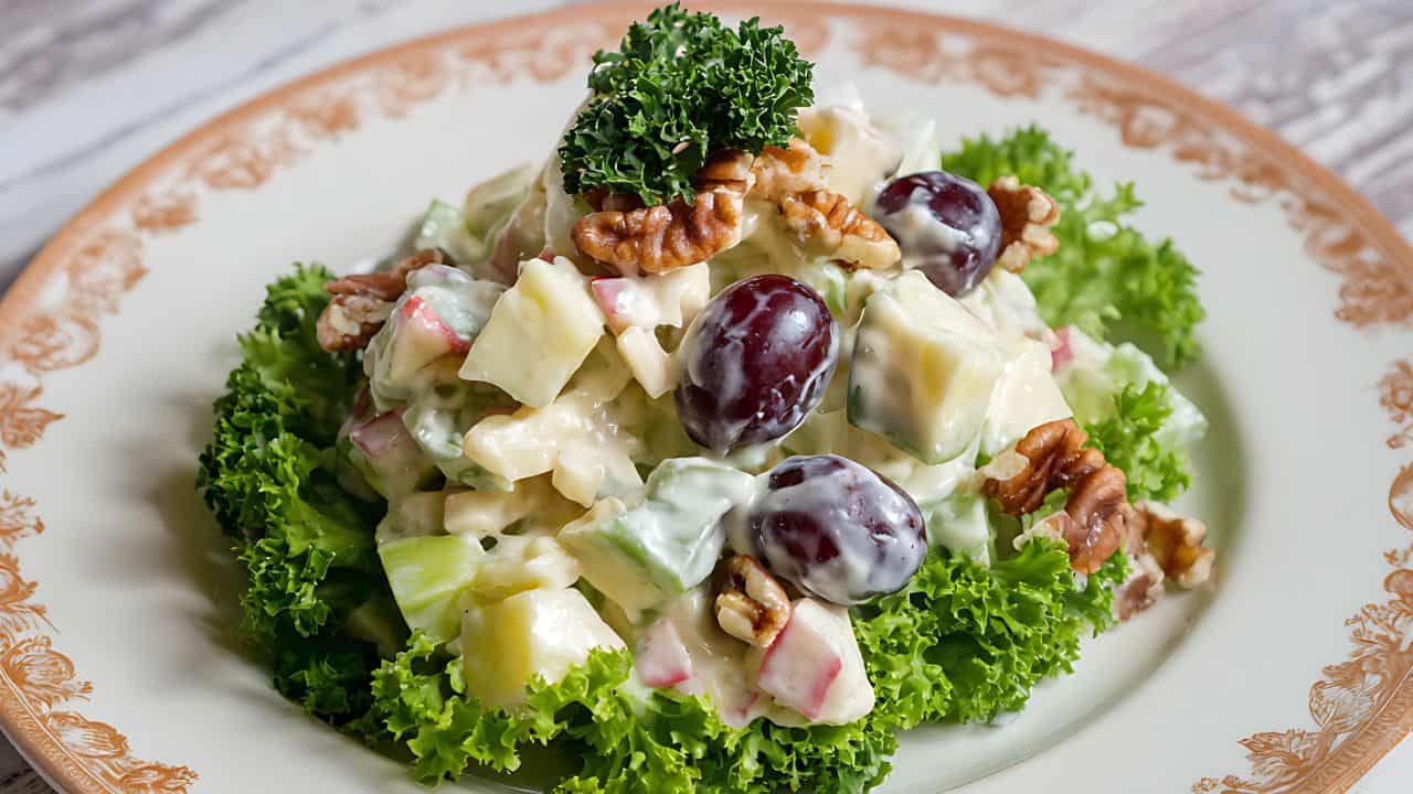 A plate of Waldorf salad with lettuce, grapes, apple, celery, walnuts, and mayonnaise dressing, garnished with a sprig of parsley on a decorative plate.