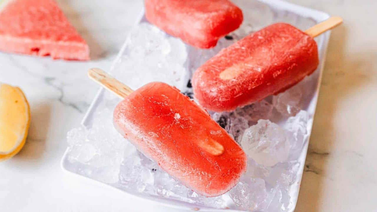 Three red popsicles on wooden sticks rest on a tray filled with ice cubes. A slice of watermelon and a lemon wedge are visible in the background on a white surface.