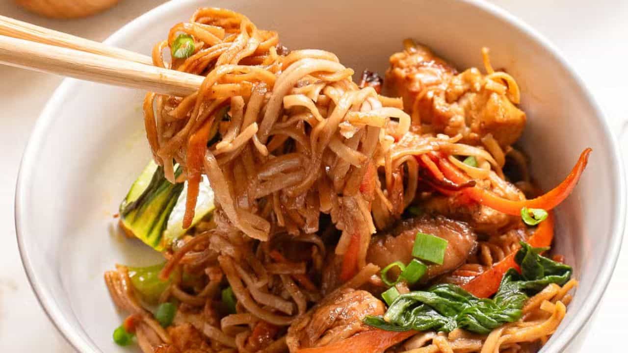 A fork lifts noodles from a white bowl filled with stir-fried noodles and vegetables. A skillet with more noodles is in the background on a marble surface.