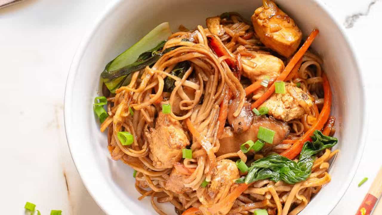 A fork lifts noodles from a white bowl filled with stir-fried noodles and vegetables. A skillet with more noodles is in the background on a marble surface.