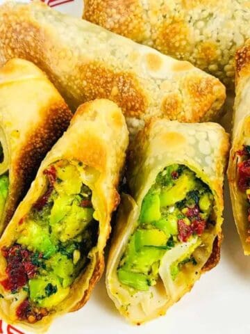 A plate with five vegetable-stuffed spring rolls, some cut open to show a green filling with herbs and spices, placed on a white plate with a red patterned border.