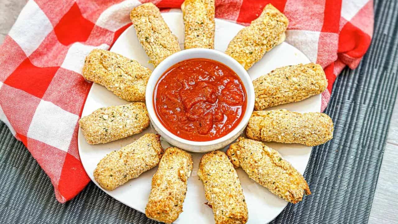 A plate of breaded mozzarella sticks arranged in a circle around a bowl of marinara sauce, with a red and white checkered cloth in the background.