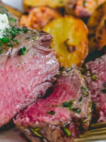 A plate of medium-rare steak topped with herb butter, served with roasted potatoes. A fork rests on the plate, and one piece of steak is sliced.