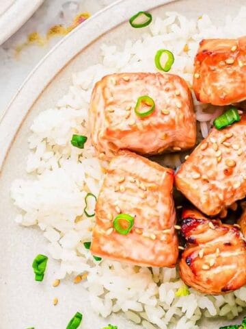 Plate of white rice topped with glazed salmon chunks, garnished with chopped green onions, served with broccoli on the side.