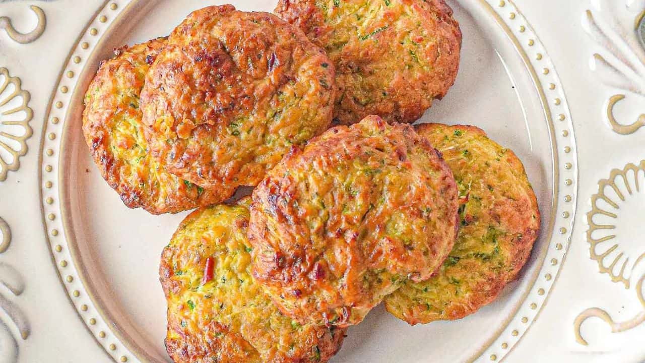 Six golden-brown zucchini fritters arranged on a white decorative plate.