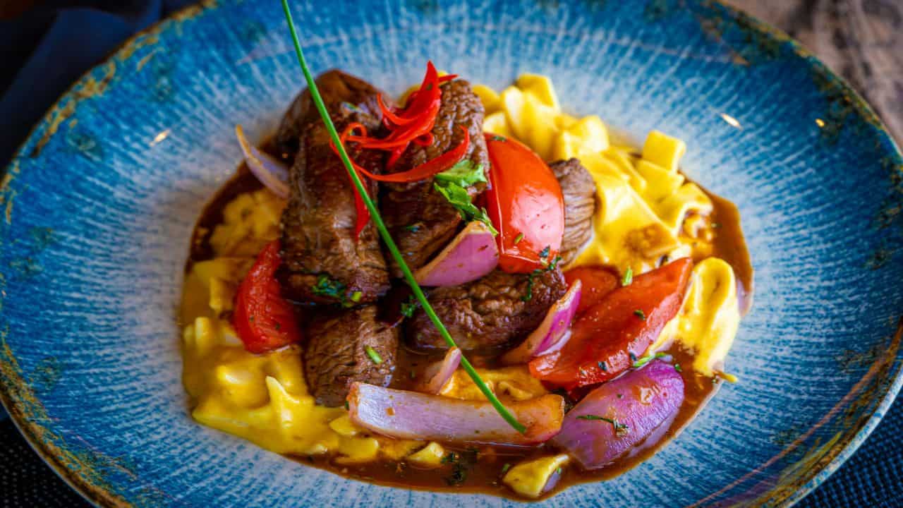 A dish of pasta topped with pieces of beef, red onion, tomato, and chili peppers, garnished with a chive, served on a blue-rimmed plate.