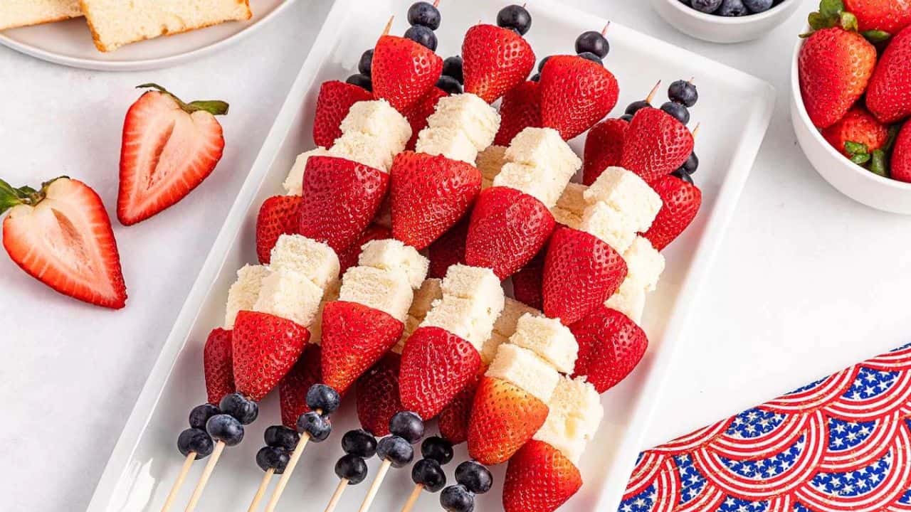Skewers with blueberries, strawberries, and cubes of white cake are arranged on a rectangular white plate, with extra fruit and cake slices nearby.