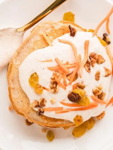 A plate of pancakes topped with white cream, shredded carrots, walnuts, and golden raisins, with a spoon and a carrot nearby.