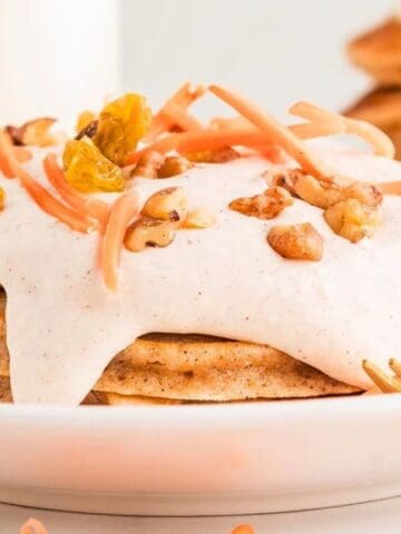 A stack of pancakes topped with cream, shredded carrots, raisins, and walnuts sits on a white plate with a gold fork.