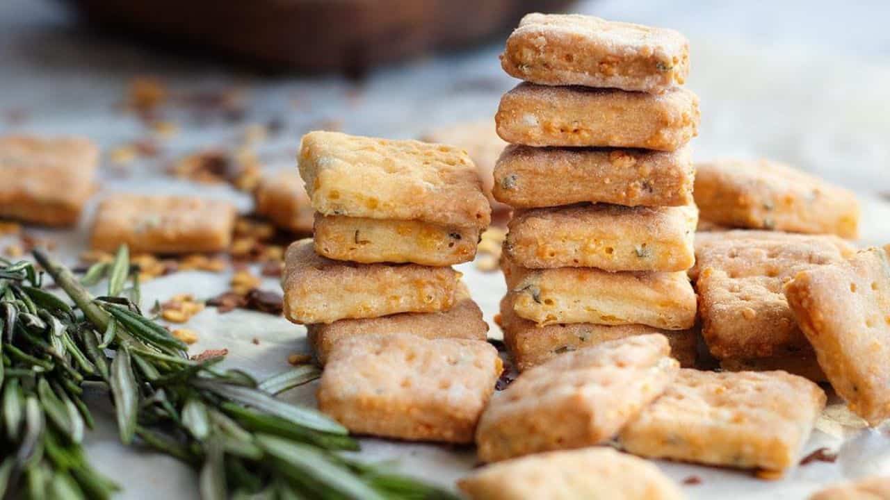 A close-up of stacked square crackers with visible herbs, scattered crackers, and a sprig of fresh rosemary on a light surface.