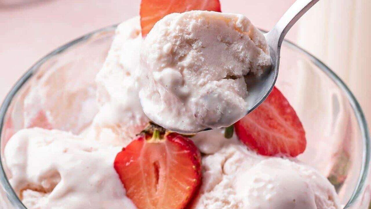 A close-up of a bowl of strawberry ice cream topped with fresh strawberry slices, with a spoon holding a scoop above the bowl.
