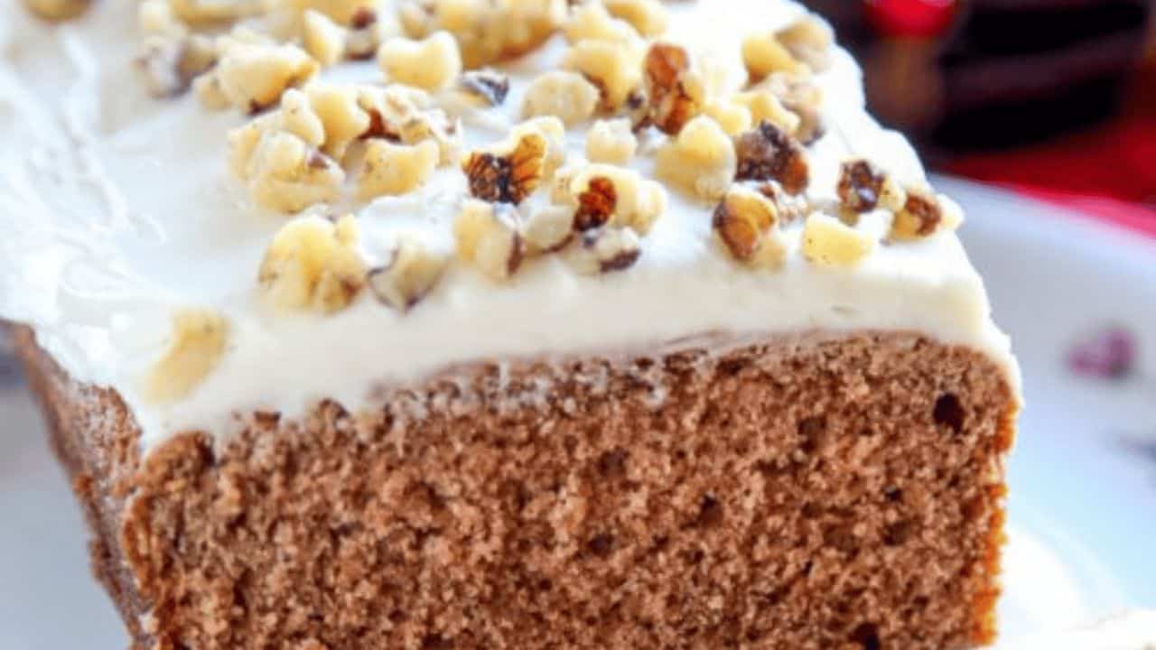 A close-up of a slice of frosted cake topped with chopped walnuts on a white plate.