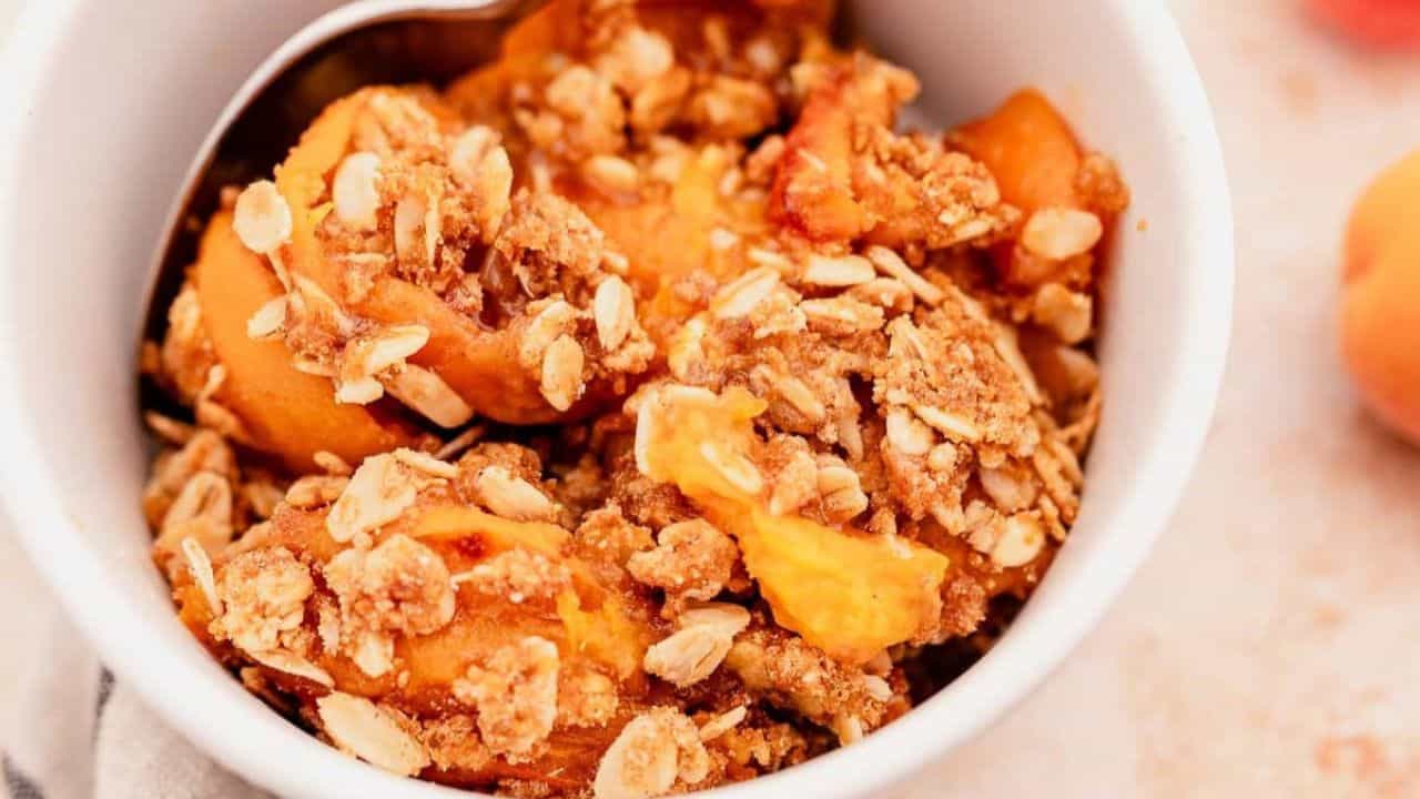 A close-up of a bowl filled with peach crisp, topped with a crumbly oat and brown sugar topping, with a spoon inside.