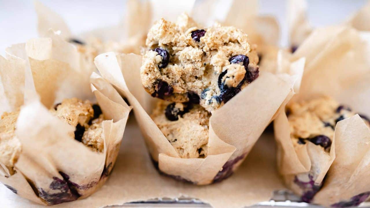 Several blueberry muffins wrapped in parchment paper are arranged closely together, with one muffin partially broken to reveal its interior texture.