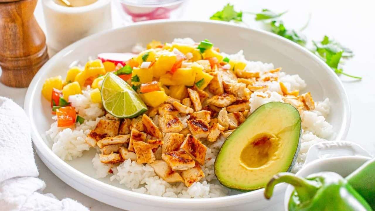 A white plate with white rice, grilled chicken pieces, diced mango salsa, half an avocado, and a lime wedge, garnished with herbs.