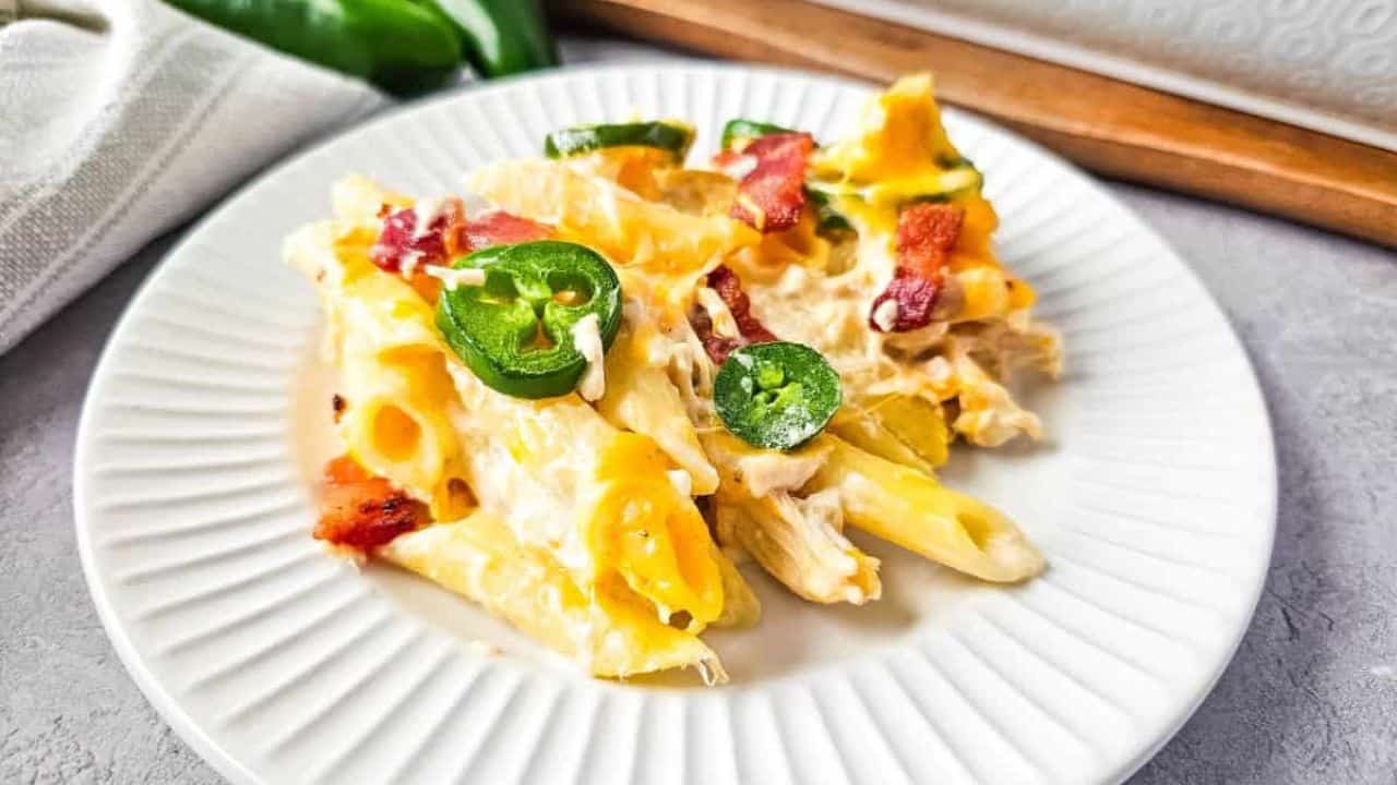 A serving spoon scoops into a baked casserole with penne pasta, cheese, jalapeños, and bacon pieces.