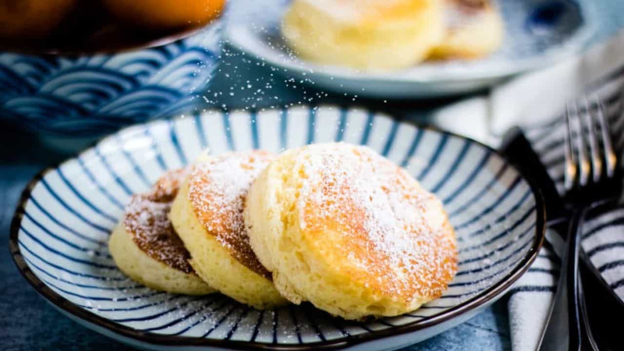 Four round Japanese souffle pancakes dusted with powdered sugar are arranged on a striped plate, with additional pastries on a patterned plate nearby. Persimmons are visible in the upper left corner.