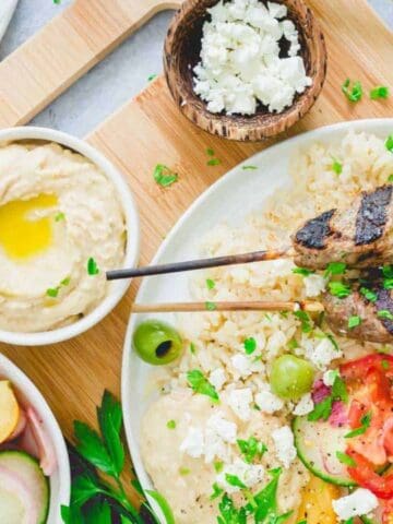 A plate with grilled meat skewers, rice, hummus, olives, feta cheese, and a salad with sliced tomatoes and cucumbers. Other bowls with hummus, olives, and salad are nearby.