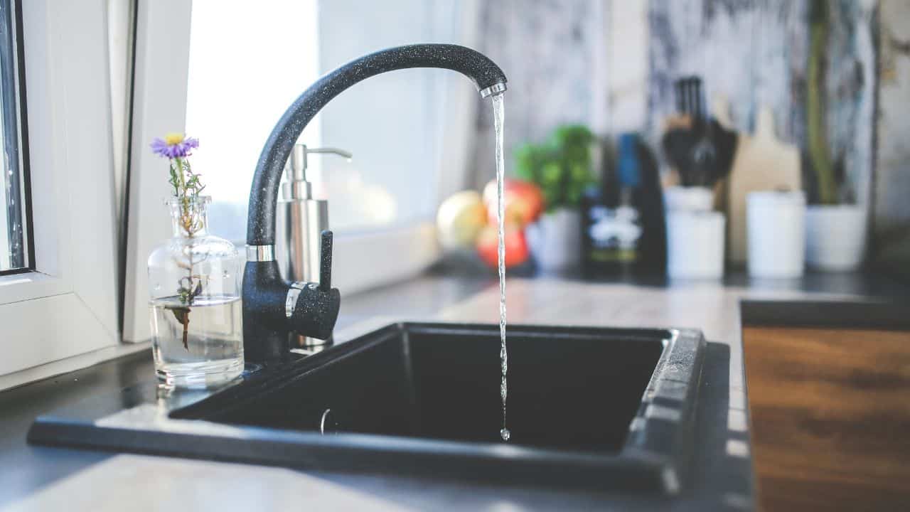 A black kitchen faucet with water running into a black sink, with a glass vase, soap dispenser, and blurred kitchen items in the background.