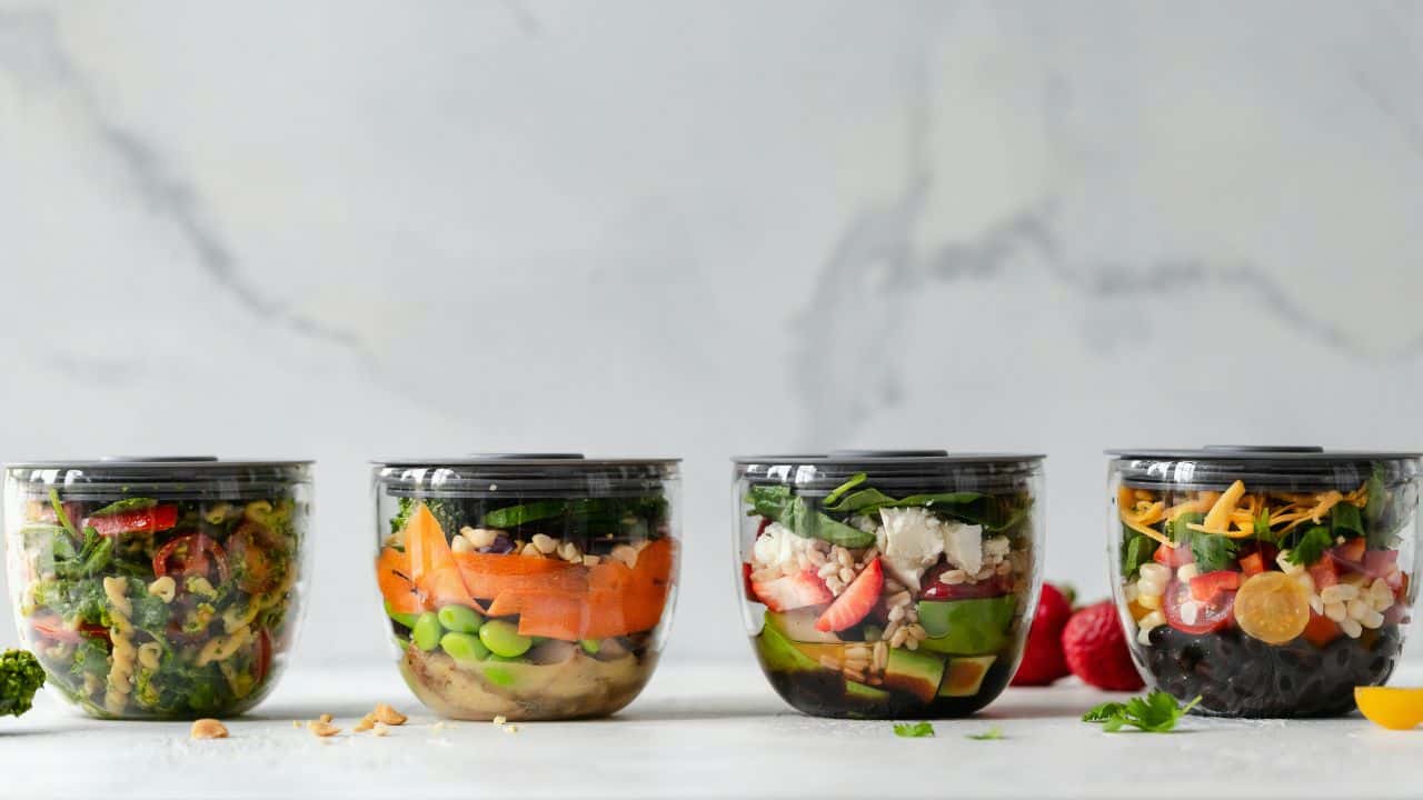 Four clear plastic containers filled with assorted layered salads, each with different colorful vegetables and toppings, arranged in a row on a light surface.