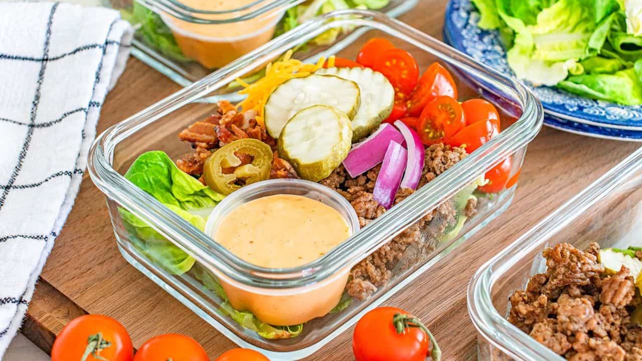 A glass meal prep container filled with lettuce, ground beef, cherry tomatoes, red onion, cheddar cheese, pickles, bacon bits, and a small cup of creamy dressing.