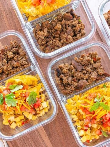 Glass meal prep containers filled with scrambled eggs, seasoned ground meat, and toppings such as cheese, cilantro, and salsa, arranged on a wooden surface with bowls of eggs and salsa nearby.