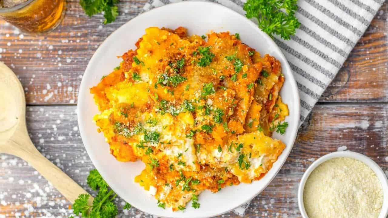 Plate of baked lasagna topped with melted cheese and chopped parsley, set on a wooden table beside a bowl of grated cheese, a striped napkin, and a wooden spoon.