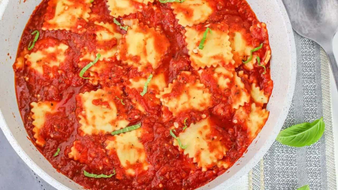 A plate of ravioli topped with red tomato sauce and garnished with fresh basil, placed on a gray tablecloth next to a fork and basil leaves.
