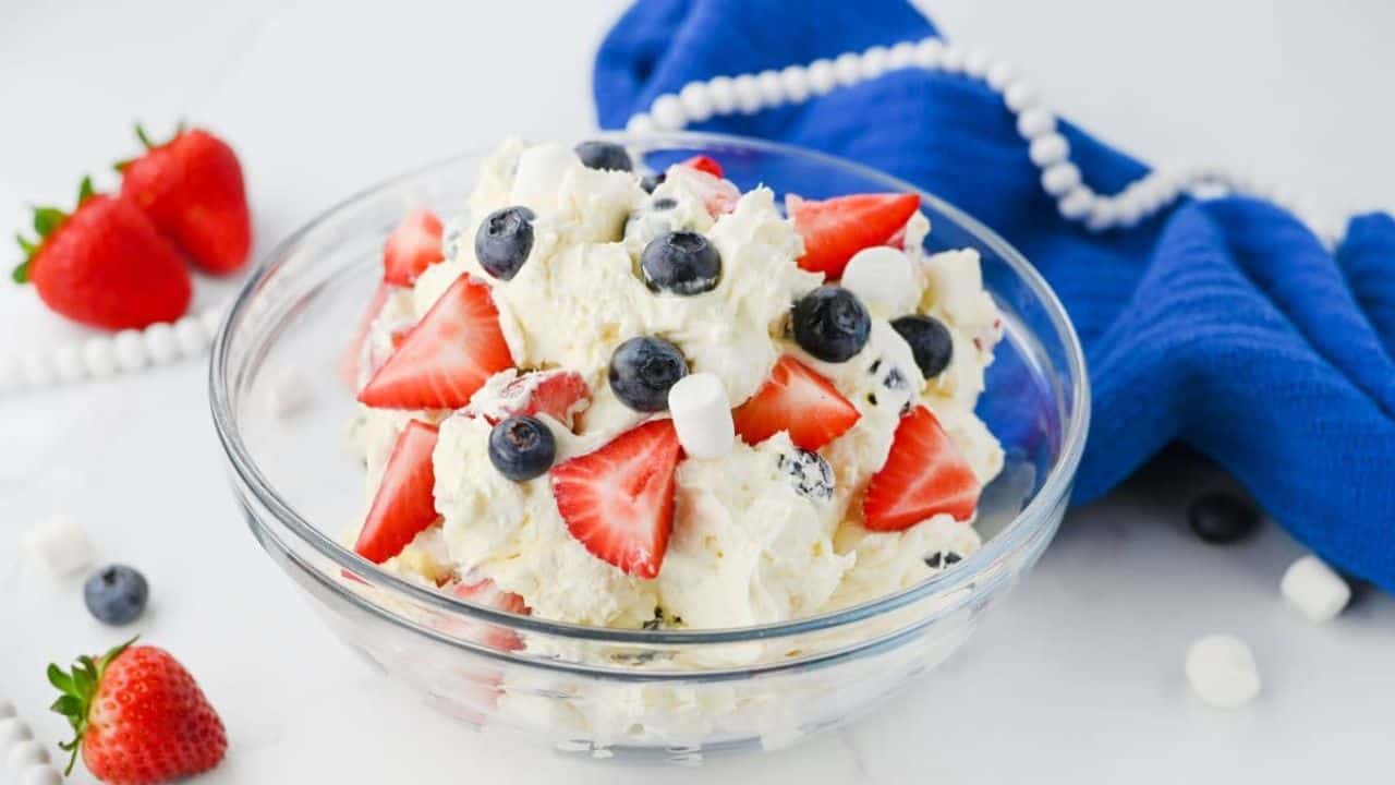 A glass bowl of patriotic dessert on a white surface, with a blue cloth and white beads in the background.
