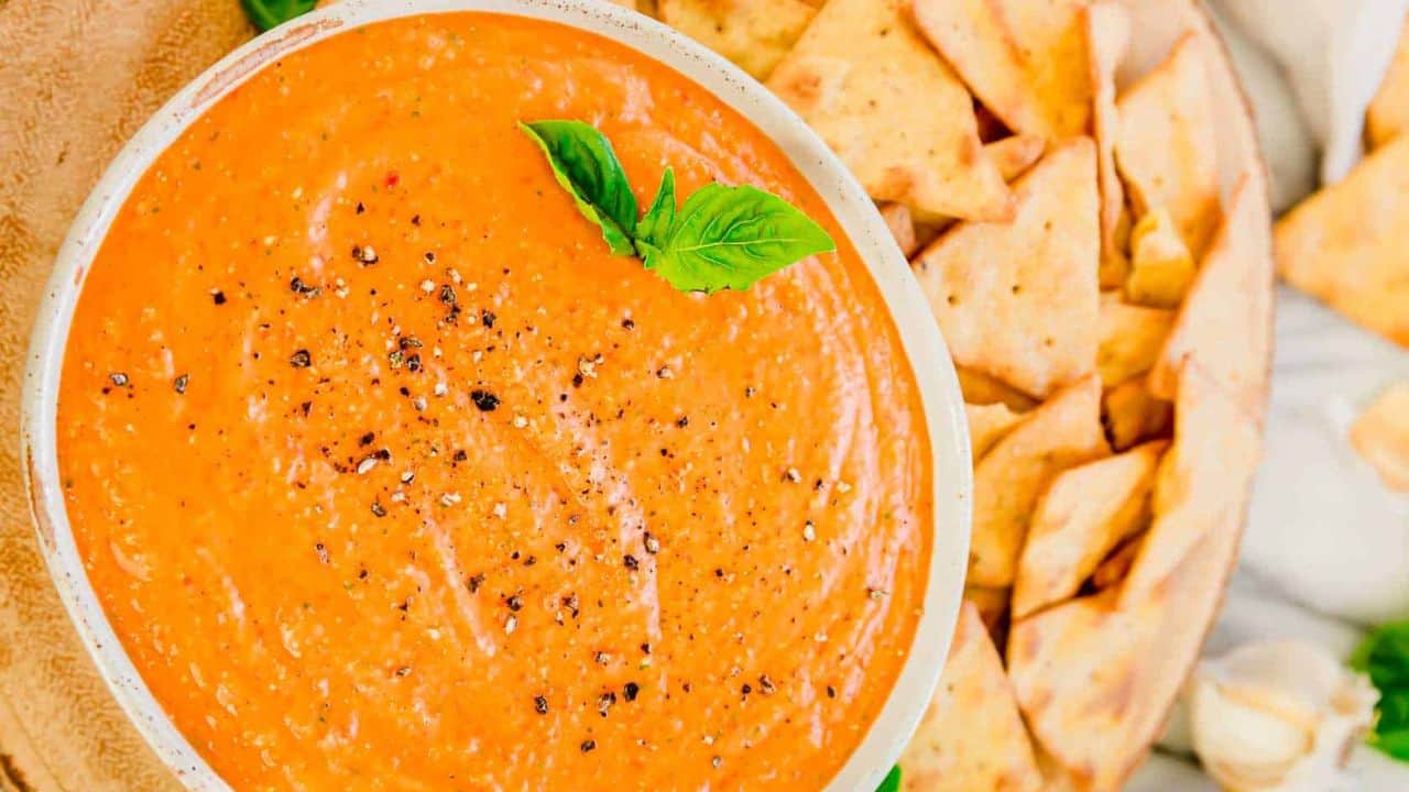 A bowl of roasted red pepper dip with crackers on a marble table.