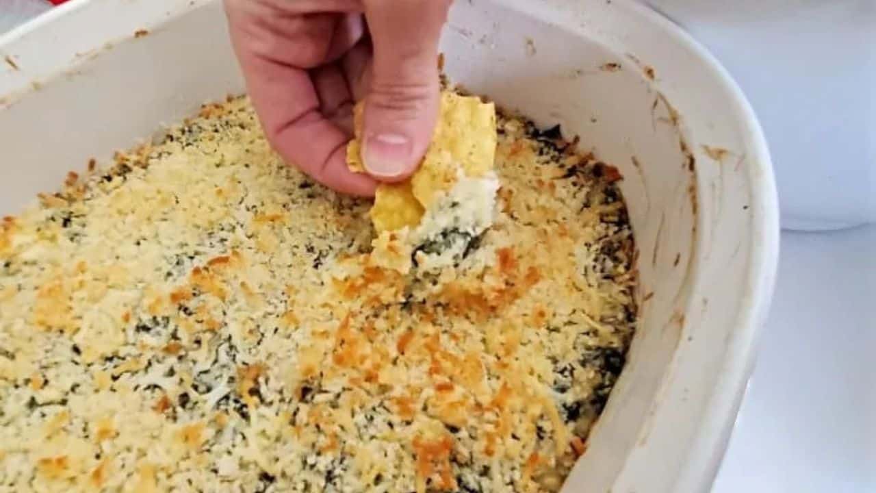 This photo shows a hand scooping out baking spinach artichoke dip from a white casserole dish.