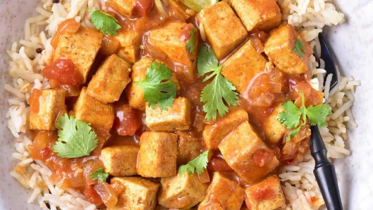 Tofu cubes coated in curry sauce with visible onions, served over fluffy rice and fresh herbs.