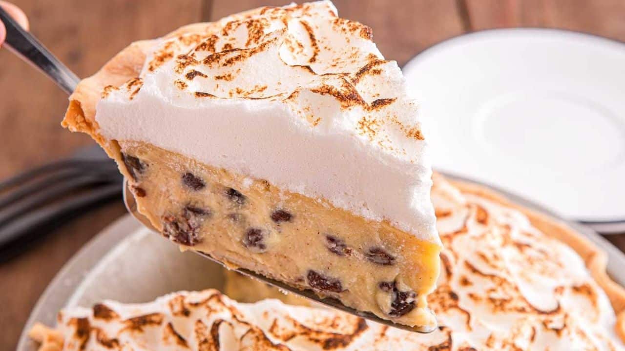 A close-up of a slice of pie with a golden-brown meringue topping and a filling containing chocolate chips, being lifted from a pie dish.