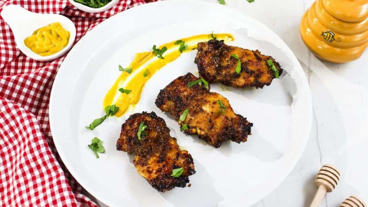 A piece of air fried chicken thigh on a fork, with a caramelized exterior and garnished with a small leaf, in focus against a blurred background of more chicken pieces.