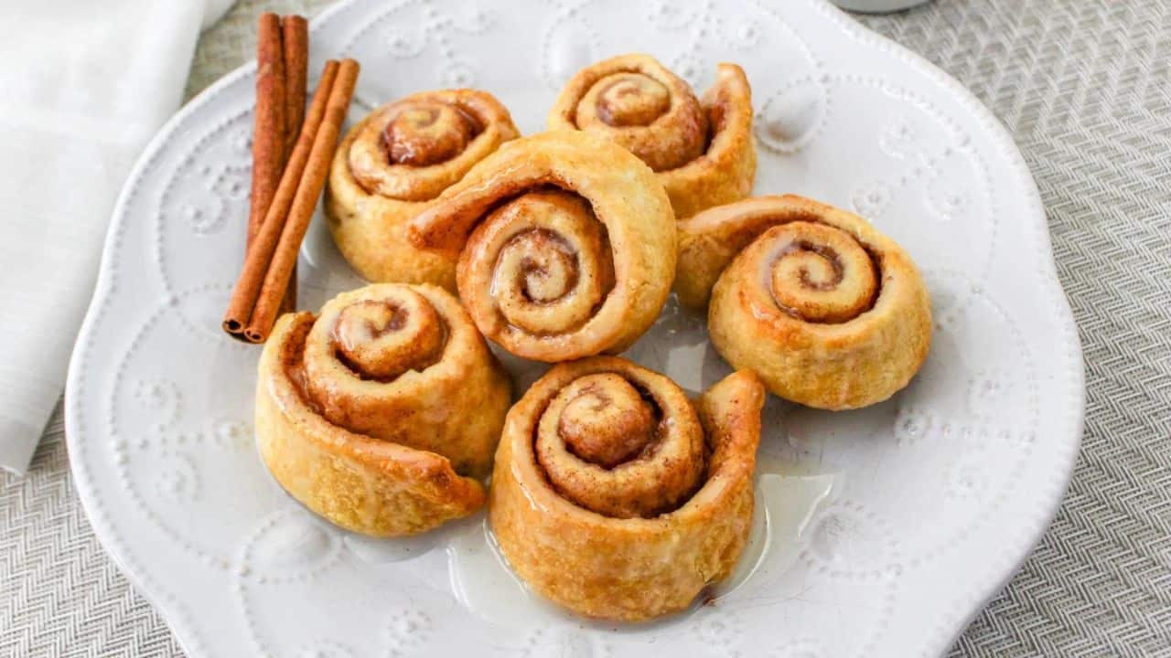 A white plate with six glazed cinnamon rolls, next to two cinnamon sticks, placed on a textured surface.