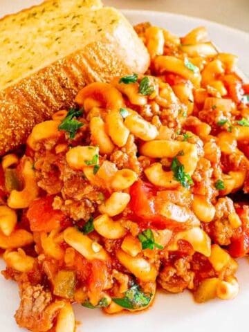 A plate of macaroni with ground meat, tomato sauce, and herbs, served with a slice of garlic bread and a fork.