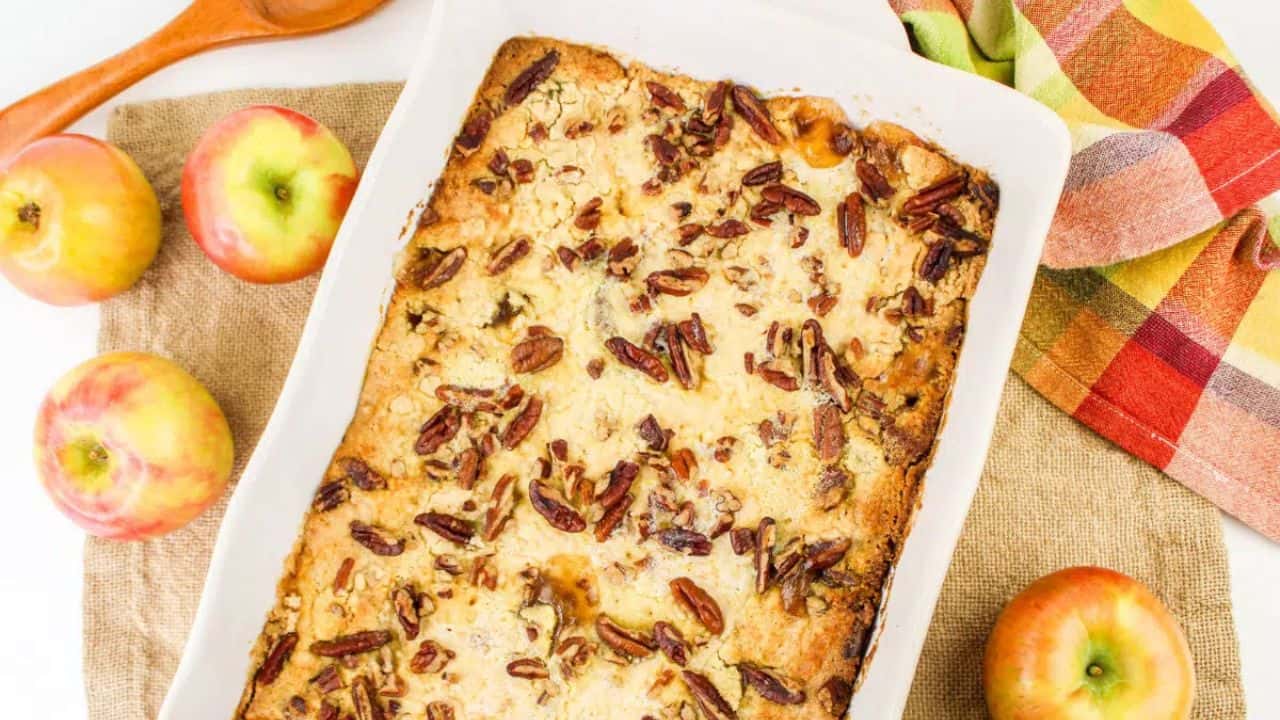 A baked apple dessert topped with pecans in a rectangular dish, surrounded by whole apples and a colorful napkin.