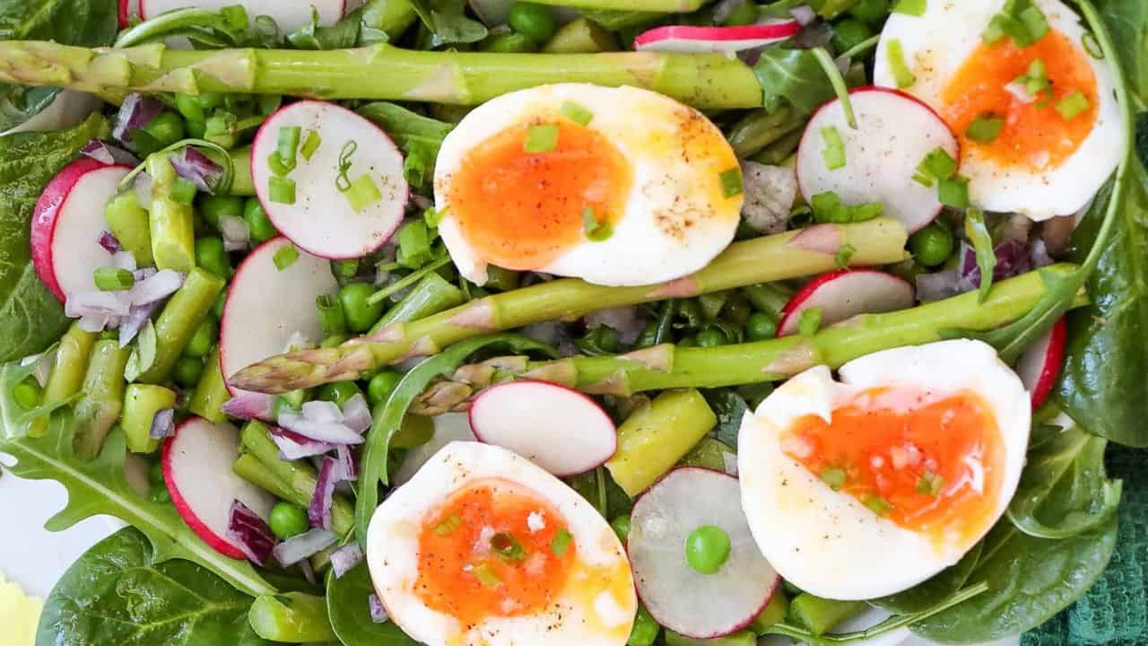 A vibrant asparagus salad with spinach, arugula, radishes, and soft-boiled eggs is topped with chopped green onions and arranged beautifully in a white bowl.