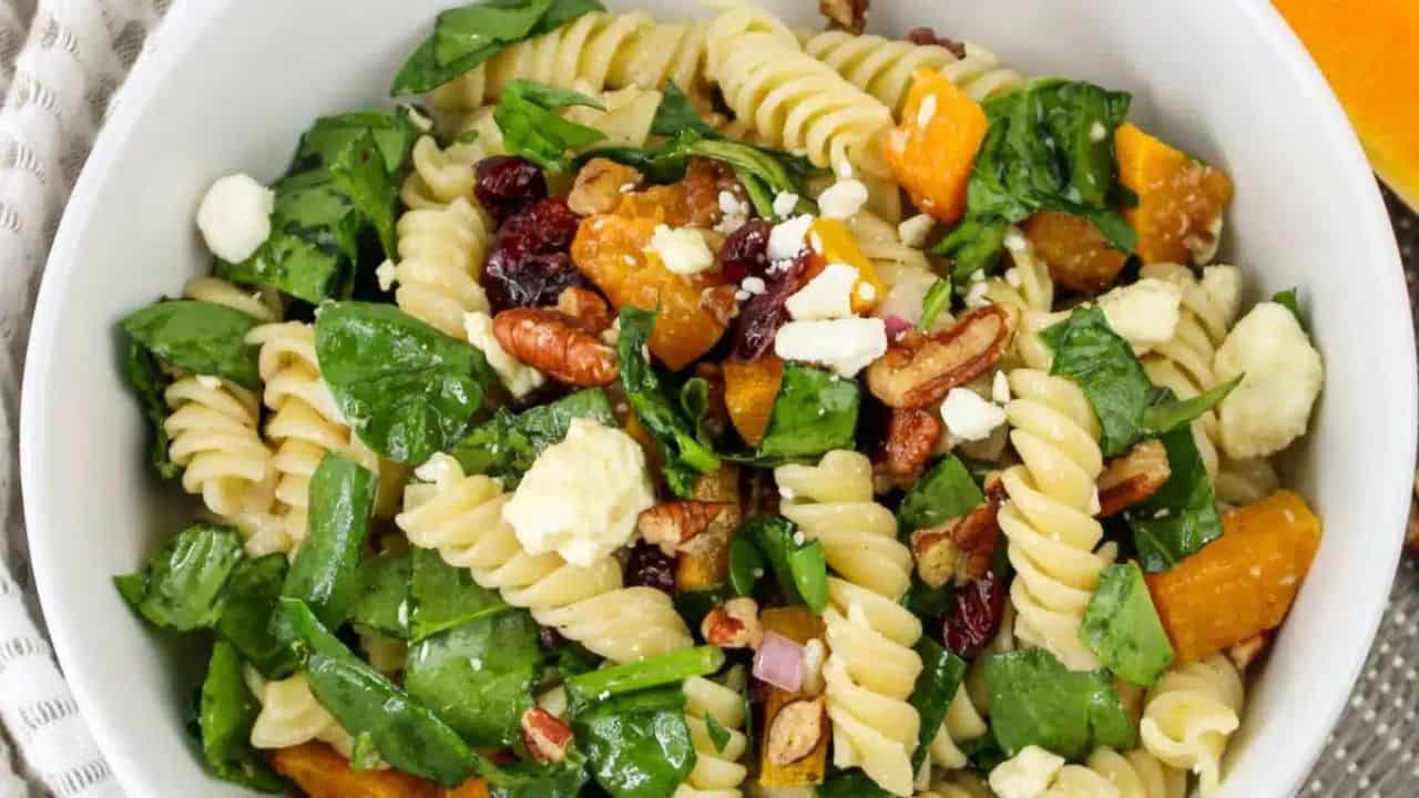 A bowl of fusilli pasta salad with spinach, roasted squash, pecans, and feta cheese, placed on a textured mat, with a spoon and sliced squash nearby.