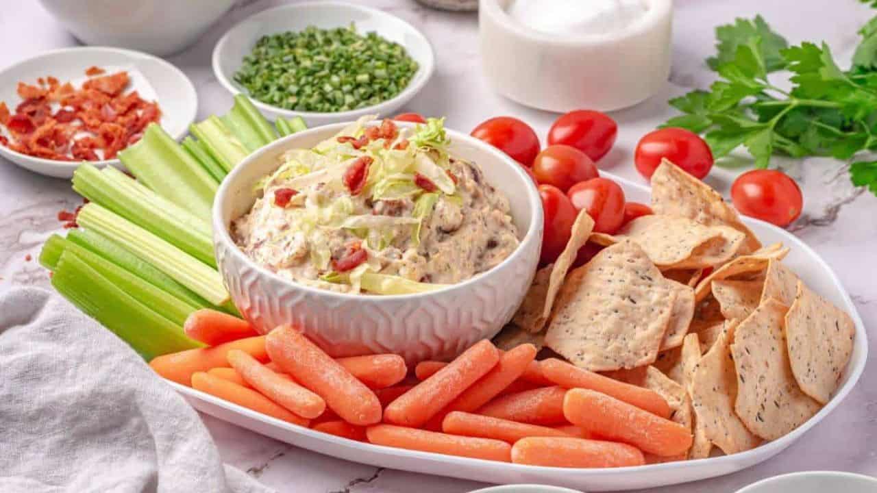A platter with a bowl of dip topped with bacon bits, surrounded by celery sticks, baby carrots, cherry tomatoes, and tortilla chips.