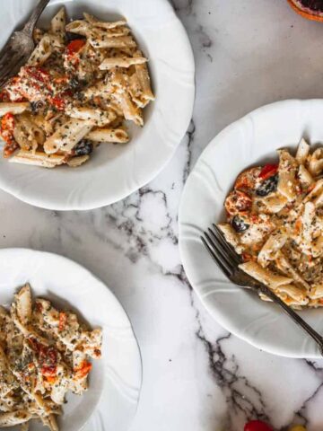 Three white plates with creamy penne pasta, tomatoes, and herbs on a marble surface, surrounded by assorted tomatoes and a casserole dish.