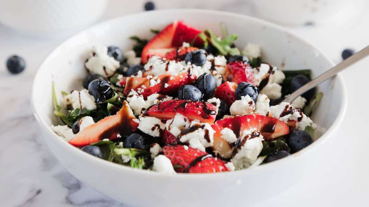 A white bowl filled with a salad of leafy greens, sliced strawberries, blueberries, crumbled white cheese, and a drizzle of dark balsamic glaze, with a metal fork on the right side.